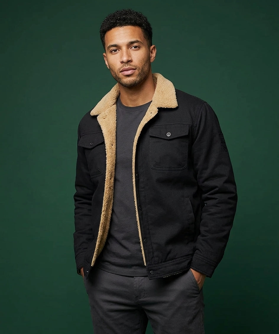 A man wearing a black jacket with a beige sherpa collar, layered over a dark gray t-shirt and dark gray pants, standing against a solid dark green background.