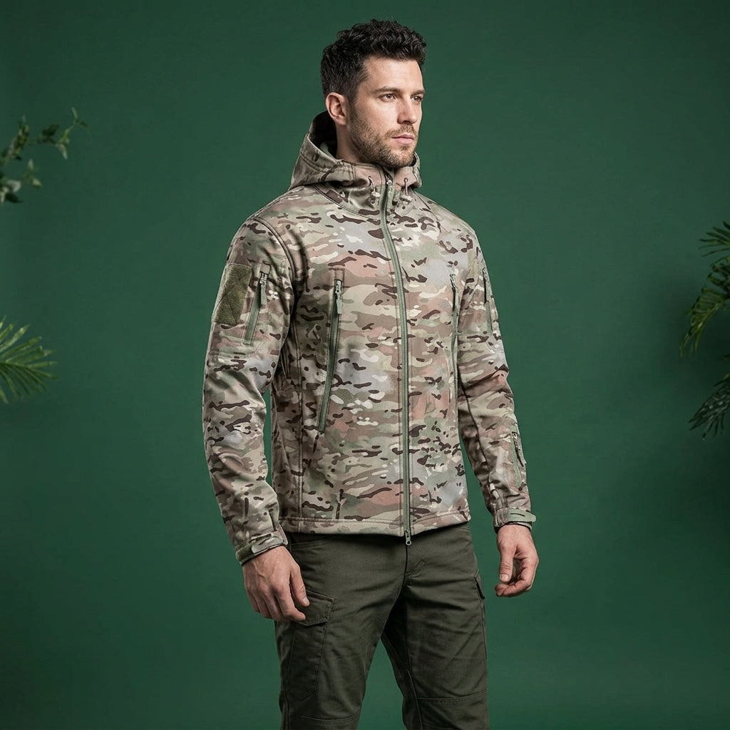 A man wearing a hooded camouflage jacket and dark olive pants, standing against a solid dark green studio background with decorative plants at the edges.