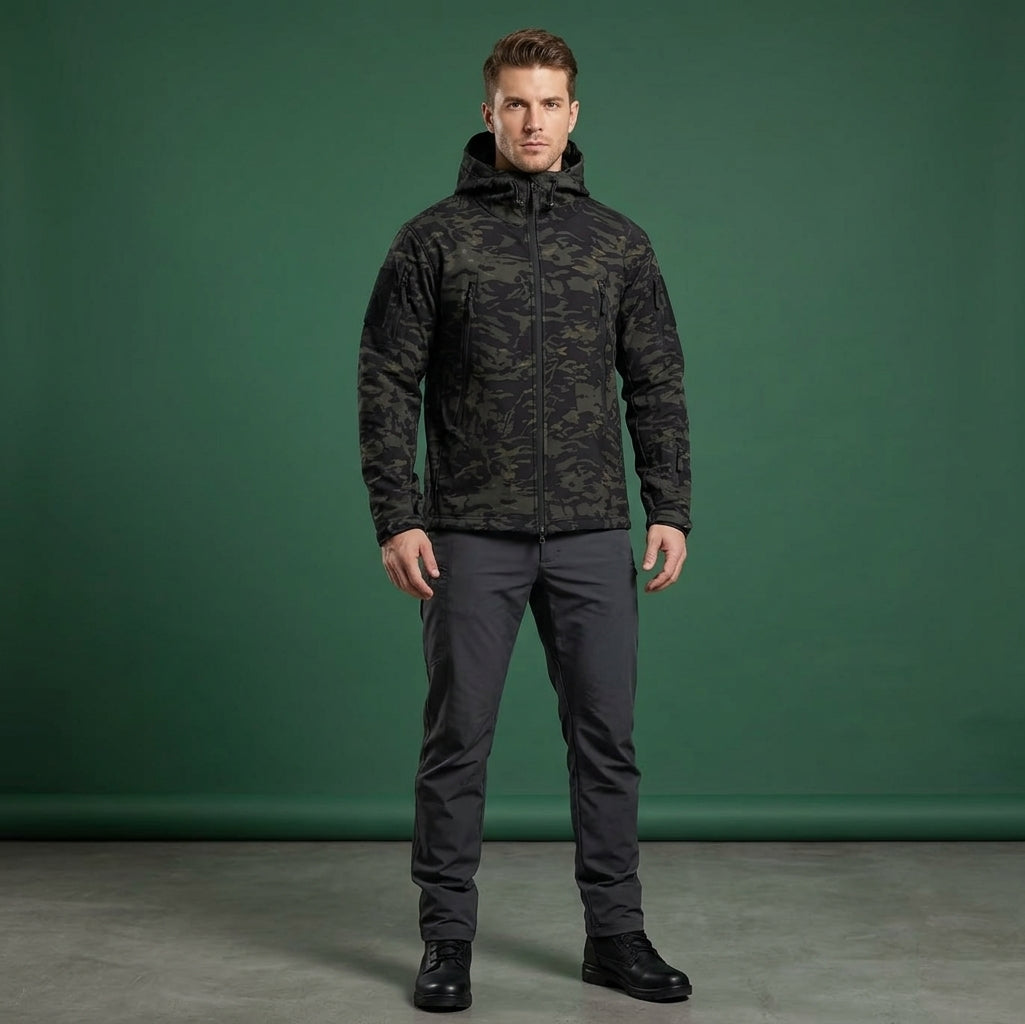 A man modeling a dark camouflage hooded jacket and dark gray pants against a solid green studio backdrop.