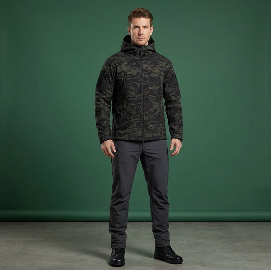 A man modeling a dark camouflage hooded jacket and dark gray pants against a solid green studio backdrop.