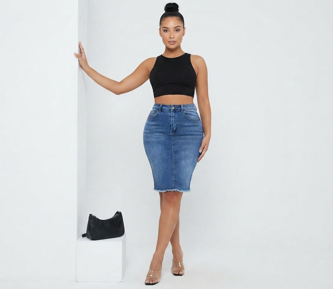 A woman modeling a black sleeveless crop top and a blue denim pencil skirt, standing in a studio setting next to a white pedestal with a small black handbag.