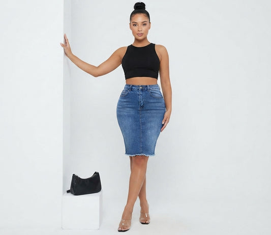 A woman modeling a black sleeveless crop top and a blue denim pencil skirt, standing in a studio setting next to a white pedestal with a small black handbag.