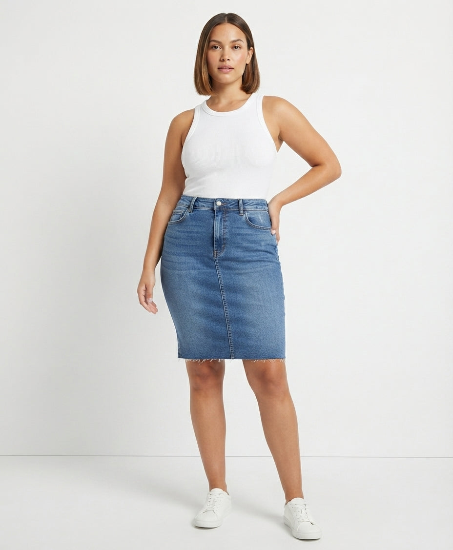 A woman modeling a blue denim pencil skirt with a frayed hem, paired with a white ribbed tank top and white sneakers, against a plain white studio background.