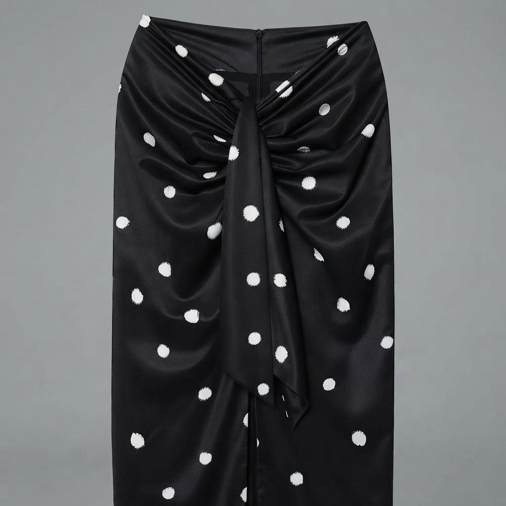 A black satin wrap skirt with white polka dots, featuring a gathered front and a central tie detail, shown against a solid gray background.
