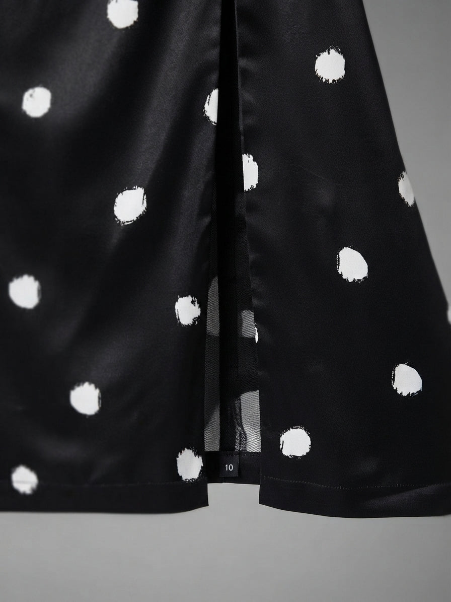 Close-up of a black satin-like garment with irregular white polka dots, showing a center seam and hem against a neutral gray background.