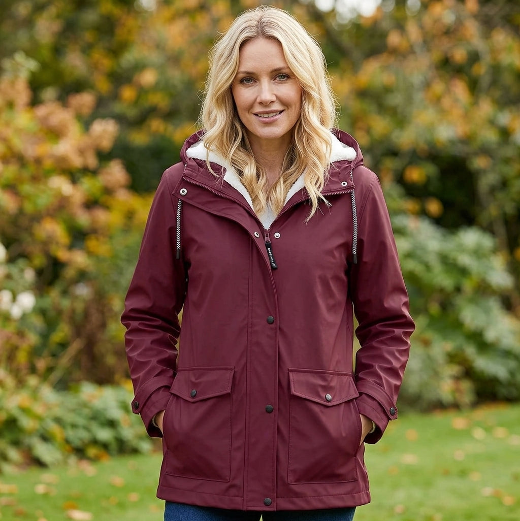 A woman wearing a maroon hooded raincoat with a white fleece-lined hood, standing outdoors in a garden setting.