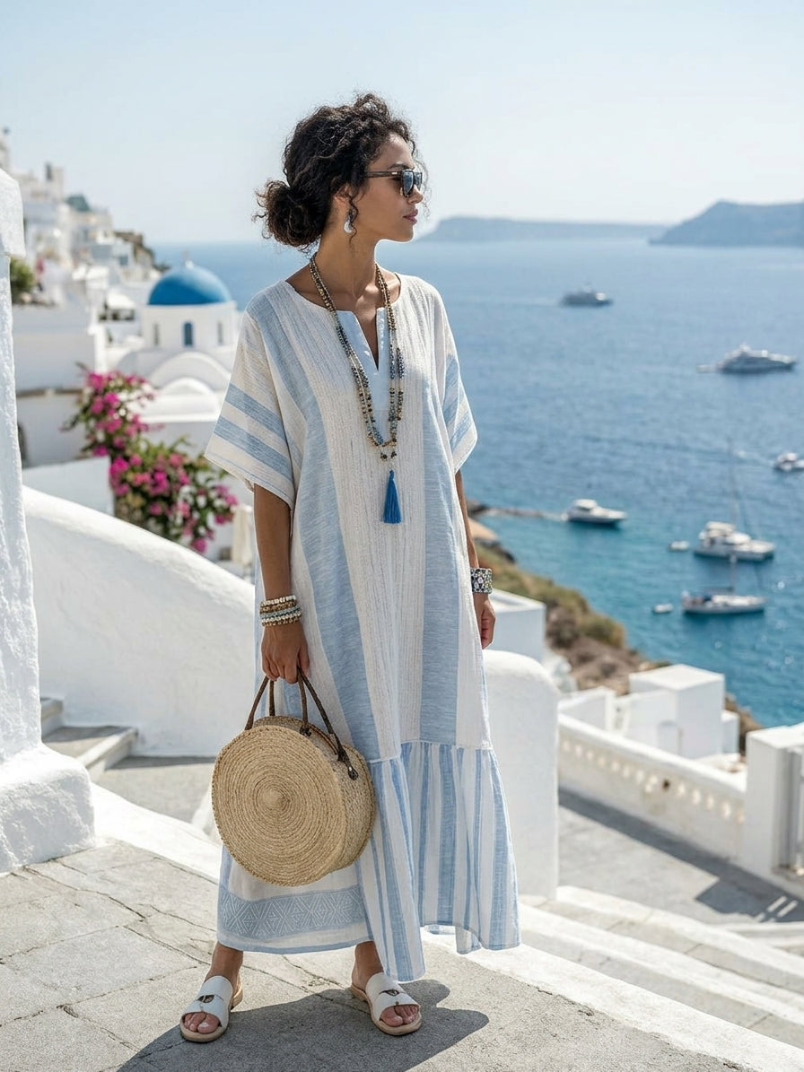 A woman wearing a light blue and white striped linen maxi dress stands on stone steps overlooking a scenic blue sea and white buildings, holding a round woven straw bag.