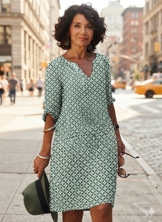 A woman stands on a city street wearing a knee-length green and white geometric patterned dress with three-quarter sleeves and a V-neckline. She is holding a dark green hat and sunglasses, with blurred city buildings and a yellow cab in the background.