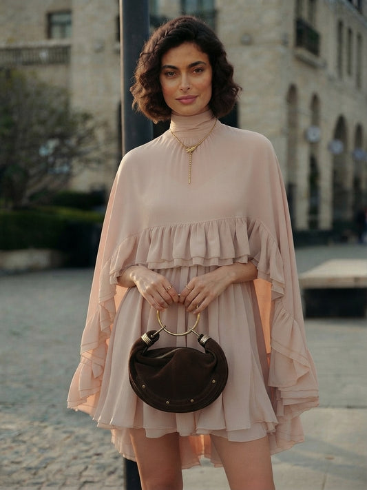 A woman wearing a pale blush pink ruffled mini dress with dramatic long sleeves, holding a dark brown crescent-shaped handbag with a gold ring handle, standing outdoors in front of a blurred stone building.