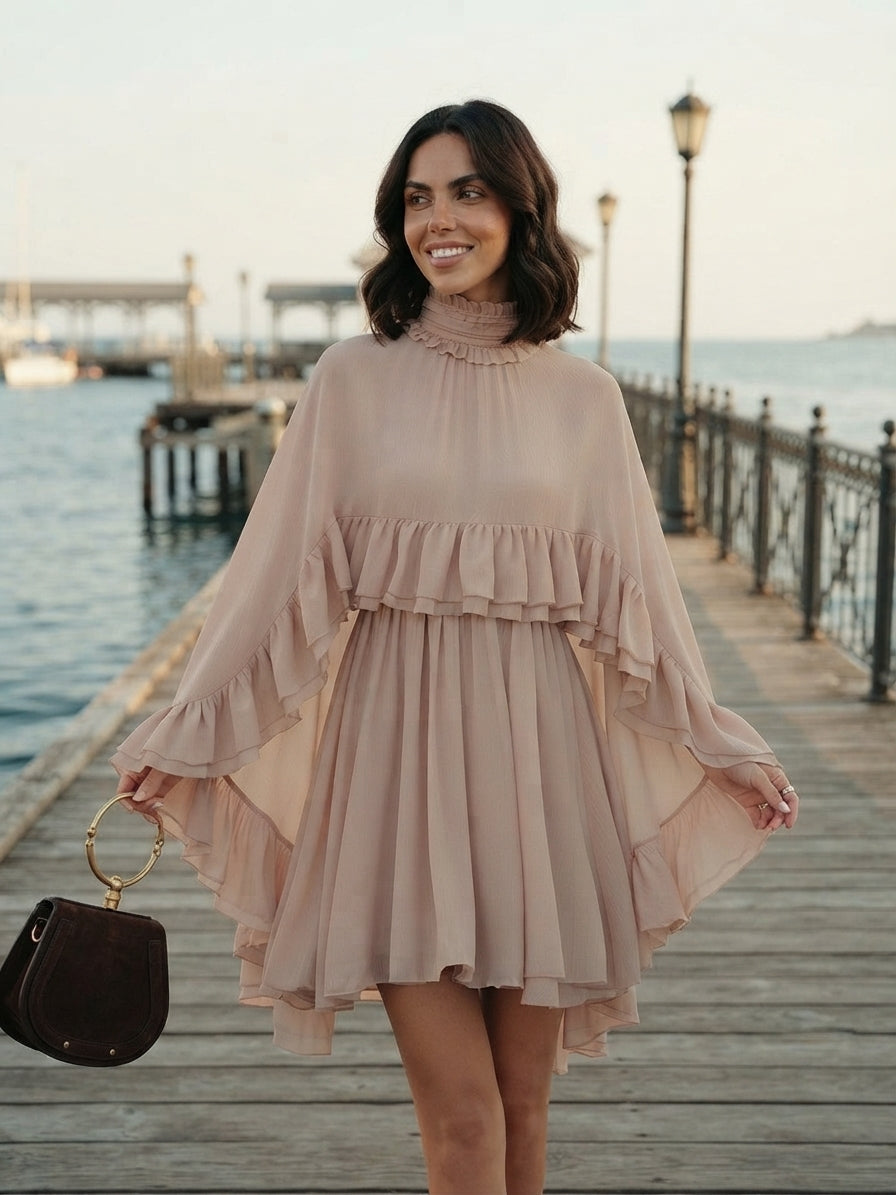 A woman stands on a wooden pier by the water wearing a light blush pink high-low mini dress with dramatic ruffled bell sleeves. She is holding a dark brown handbag with a gold ring handle.