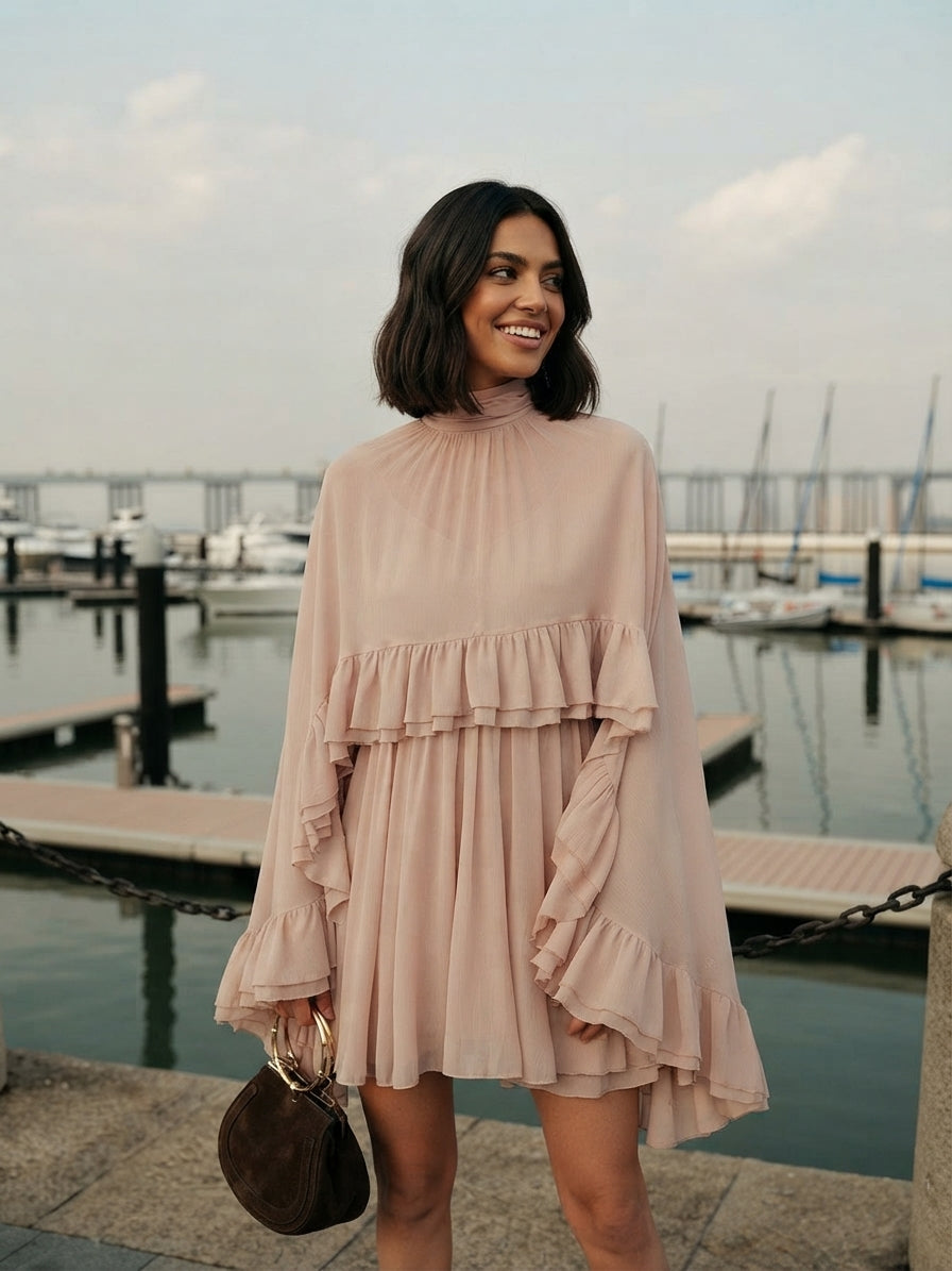 A woman wearing a light pink high-neck mini dress with layered ruffle details stands by a waterfront marina, holding a small brown handbag.