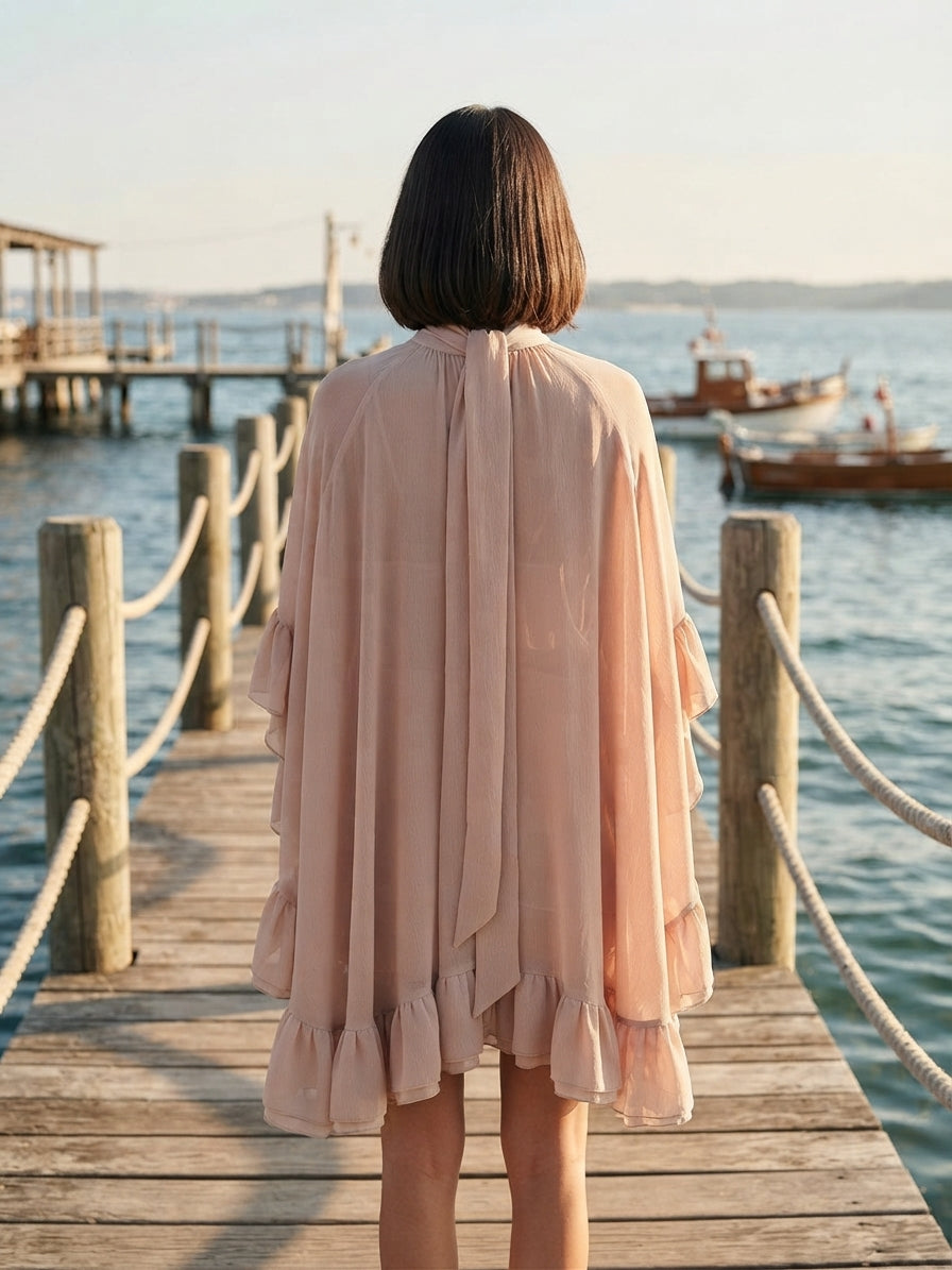 A woman viewed from behind wearing a light blush pink, flowy cover-up or tunic with ruffled edges, standing on a wooden dock overlooking a body of water with boats in the distance.