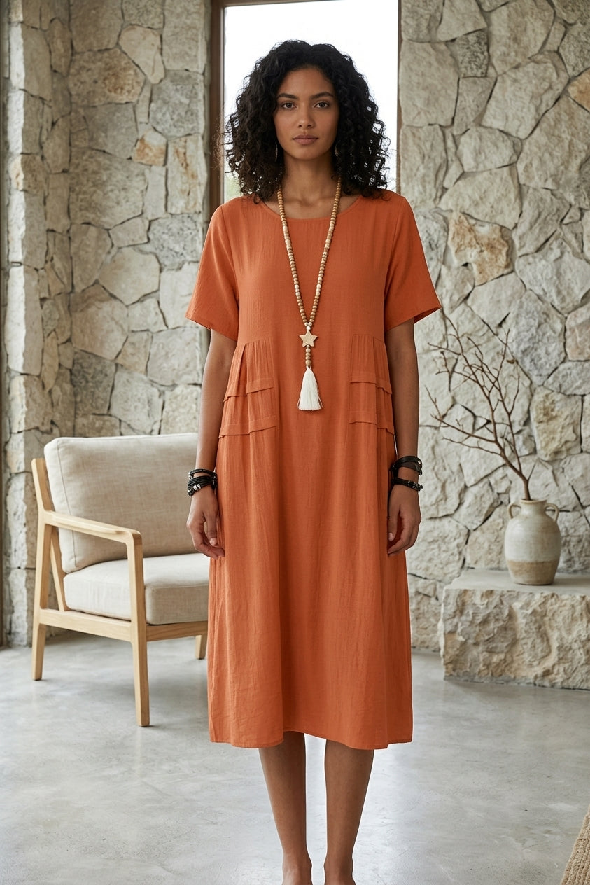 A woman wearing a short-sleeved, knee-length terracotta orange dress with a gathered waist and front pockets, standing in a rustic room with a stone wall and a light-colored armchair.
