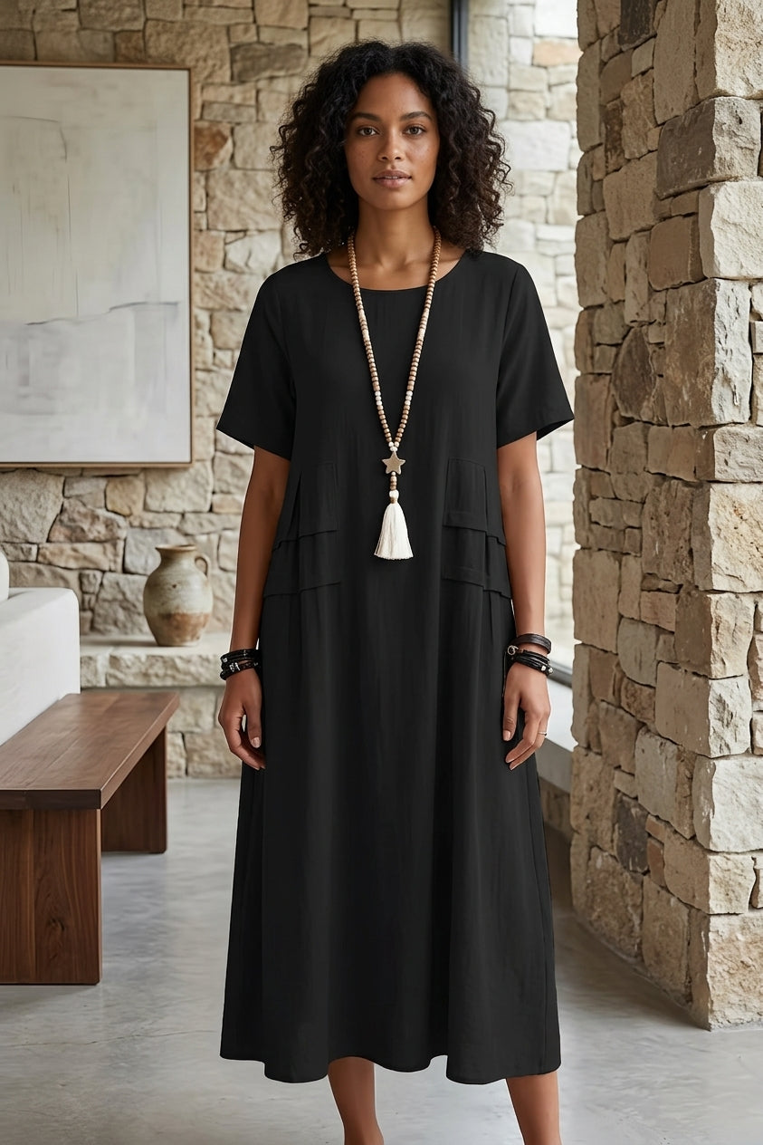 A woman wearing a black short-sleeve midi dress with a tiered waist, styled with a long white tassel necklace, standing in a rustic stone-walled interior.