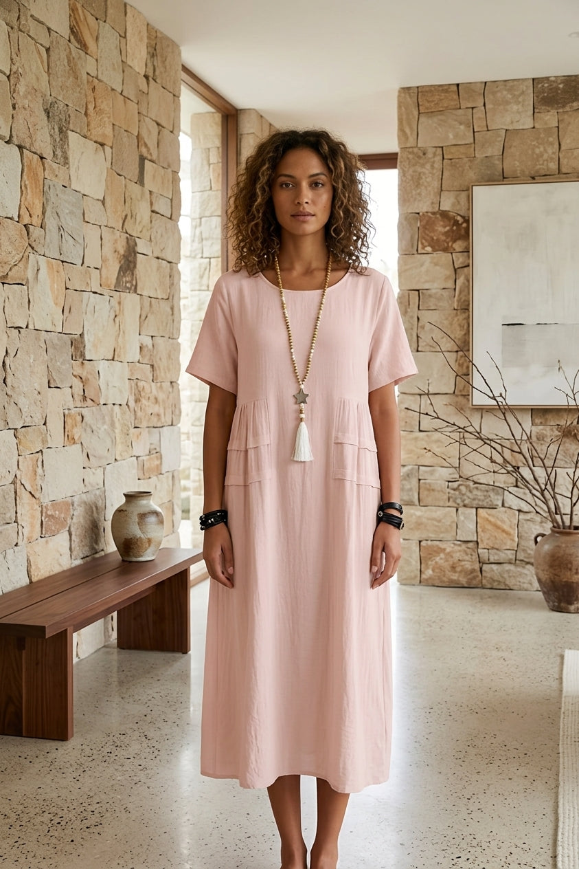 A woman stands in a stone-walled interior wearing a light pink short-sleeve midi dress with front patch pockets and a long tassel necklace.