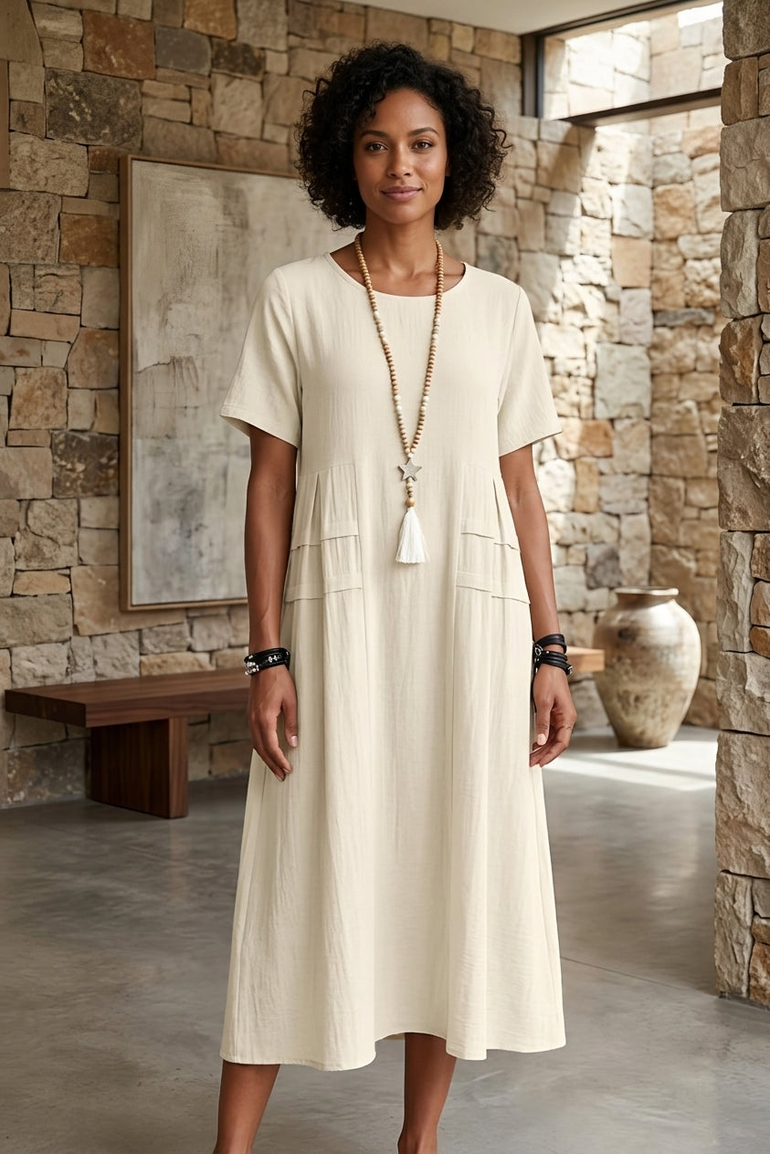 A woman wearing a short-sleeve cream-colored midi dress with a tassel necklace, standing in a rustic stone-walled interior.