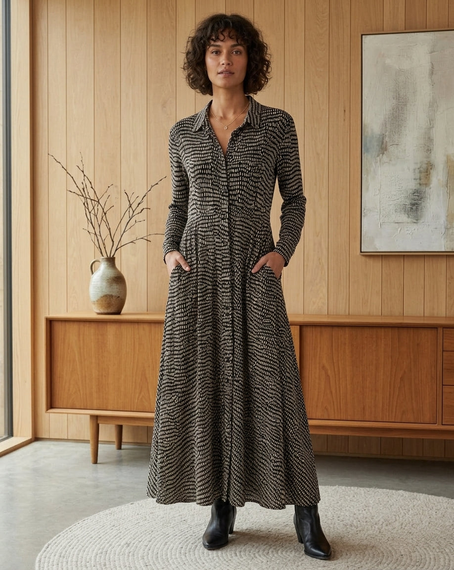 A woman stands in a modern wood-paneled room wearing a long-sleeve collared maxi dress with a black and beige wavy pattern, paired with black ankle boots.