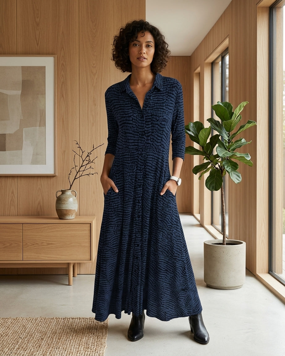 A woman stands in a modern interior wearing a long navy blue textured maxi dress with a collared neckline, front buttons, and side pockets. The room has light wood walls, a wooden sideboard, a potted plant, and large windows.
