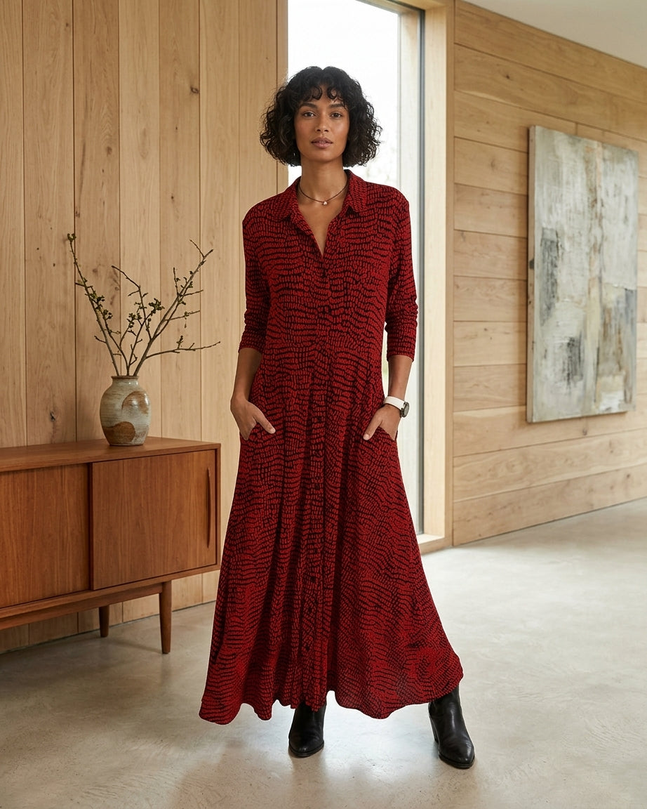 A woman wearing a long-sleeved, button-front maxi dress with a textured red and black pattern, standing in a modern room with light wood-paneled walls.