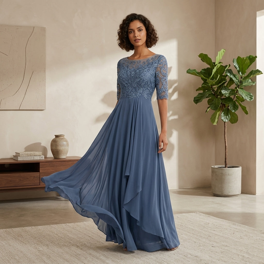 A woman models a floor-length dusty blue gown with a lace bodice, sheer elbow-length sleeves, and a flowing chiffon skirt in a warm, modern interior setting.