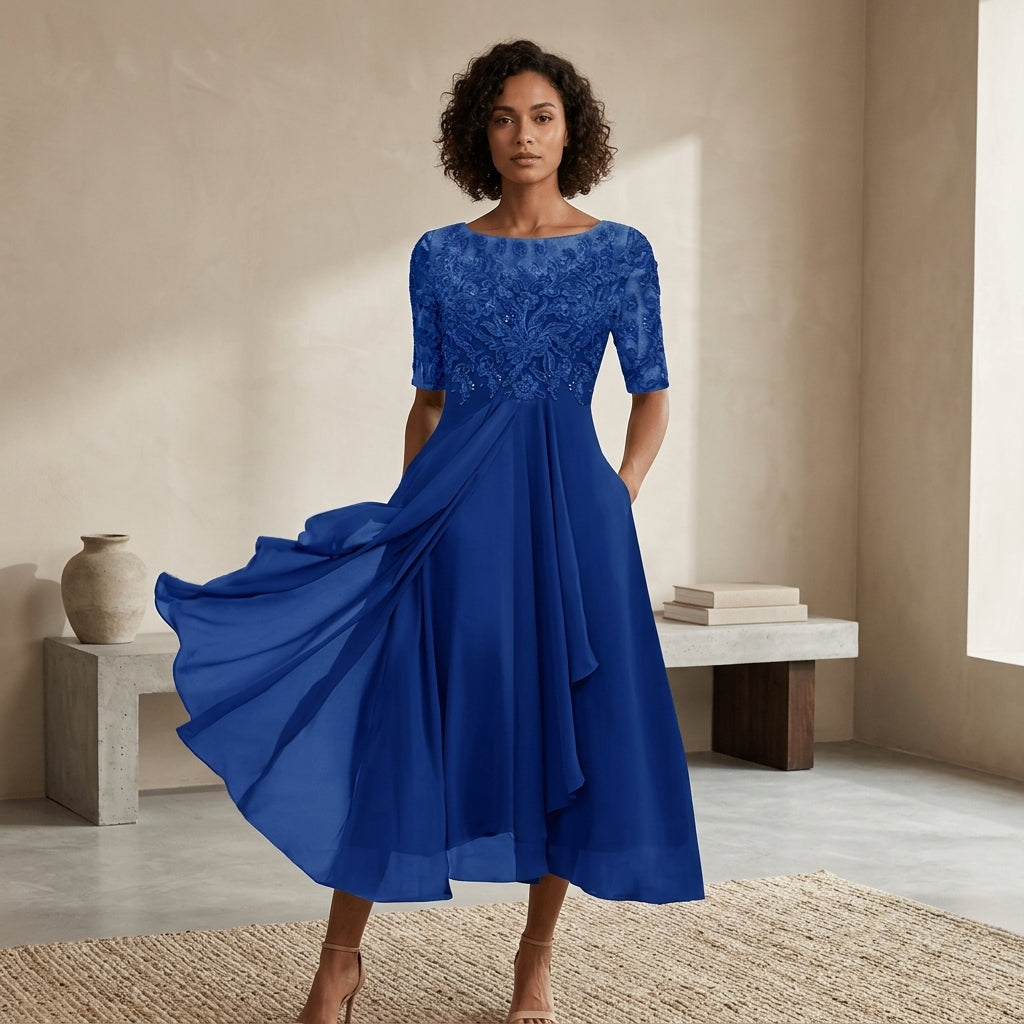 A woman models a royal blue midi dress with a lace bodice and flowing chiffon skirt in a warm, minimalist interior setting.