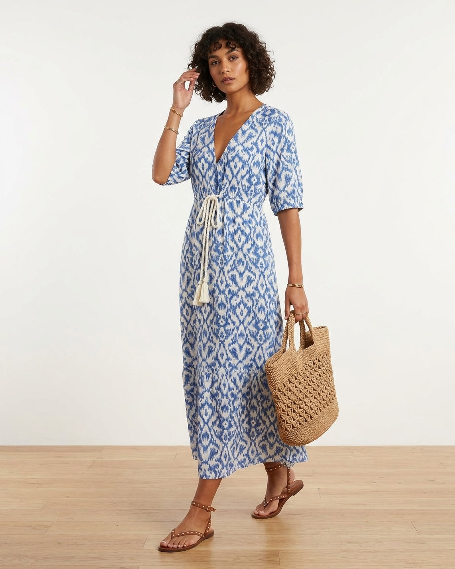 A woman modeling a blue and white patterned midi dress with a tie waist, holding a woven straw tote bag, standing on a light wood floor against a white wall.
