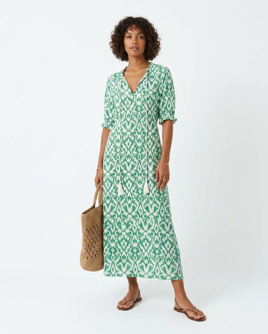 A woman modeling a green and white patterned midi dress with short sleeves, a V-neckline, and tassel ties, holding a woven straw tote bag against a plain white studio background.