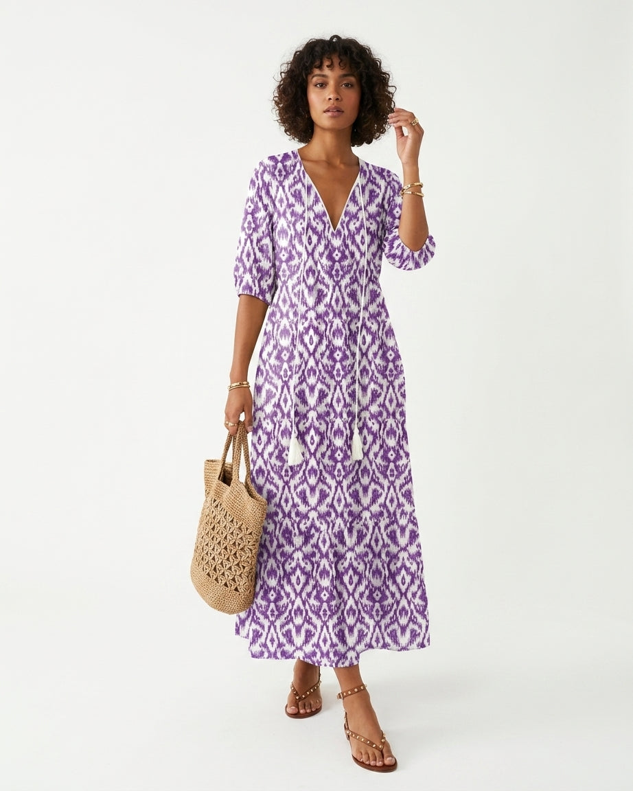 A woman modeling a purple and white patterned midi dress with three-quarter sleeves, holding a woven straw tote bag against a plain white studio background.