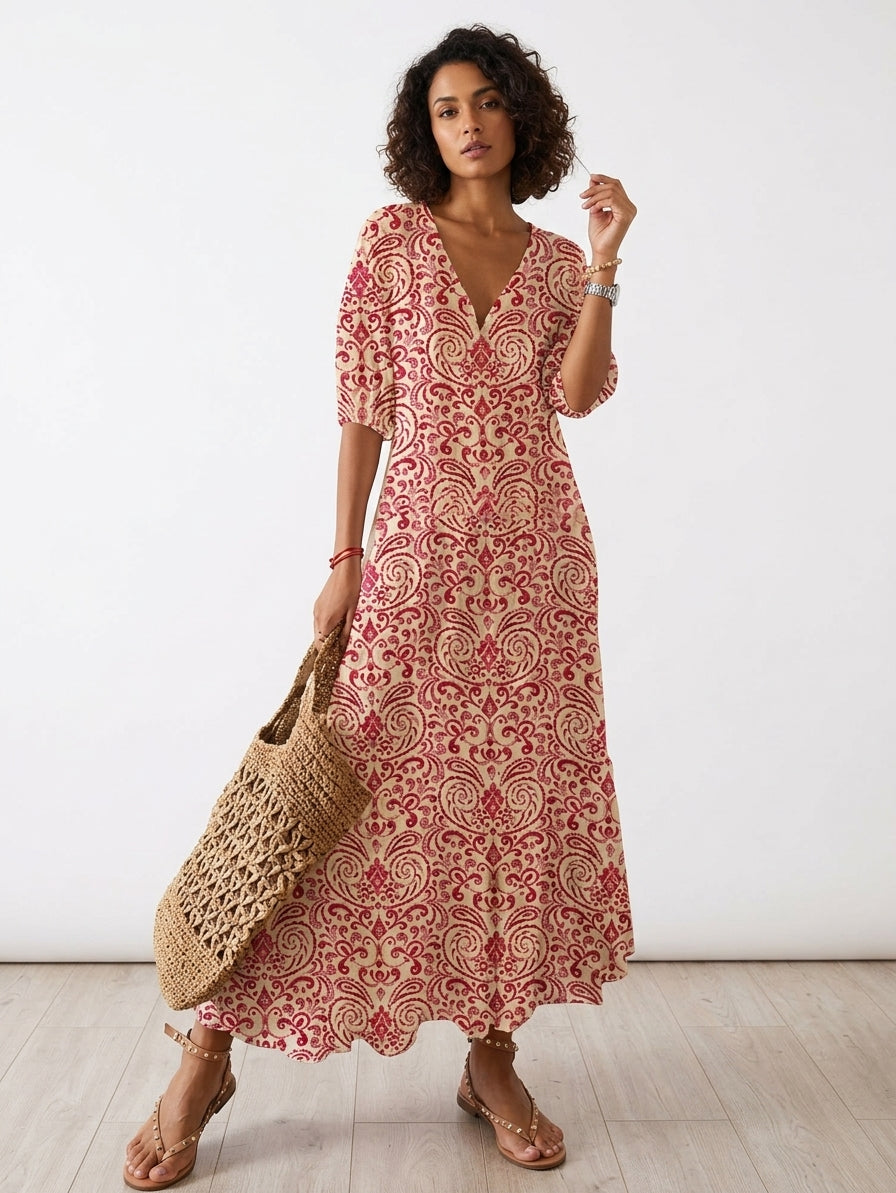 A woman modeling a bohemian-style midi dress with a red and beige paisley pattern, holding a woven straw tote bag against a white studio background.
