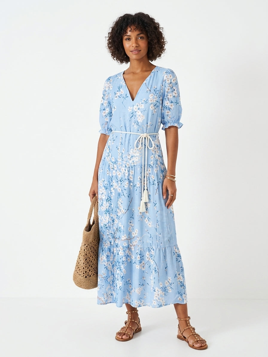 A woman modeling a light blue floral midi dress with a white tie belt, holding a woven straw tote bag against a plain white studio background.