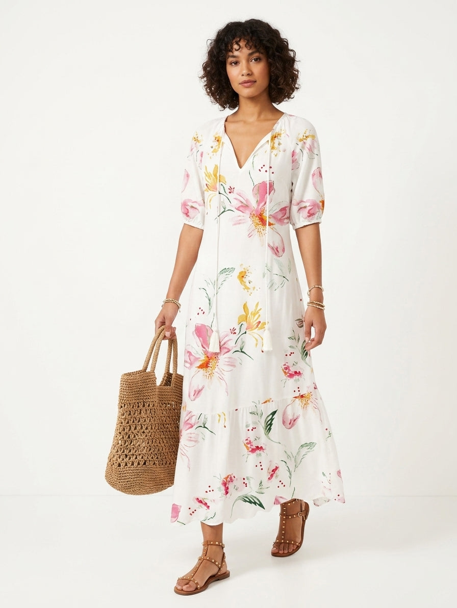 A woman modeling a white midi dress with a pink, yellow, and green floral print, holding a brown woven tote bag against a plain white studio background.