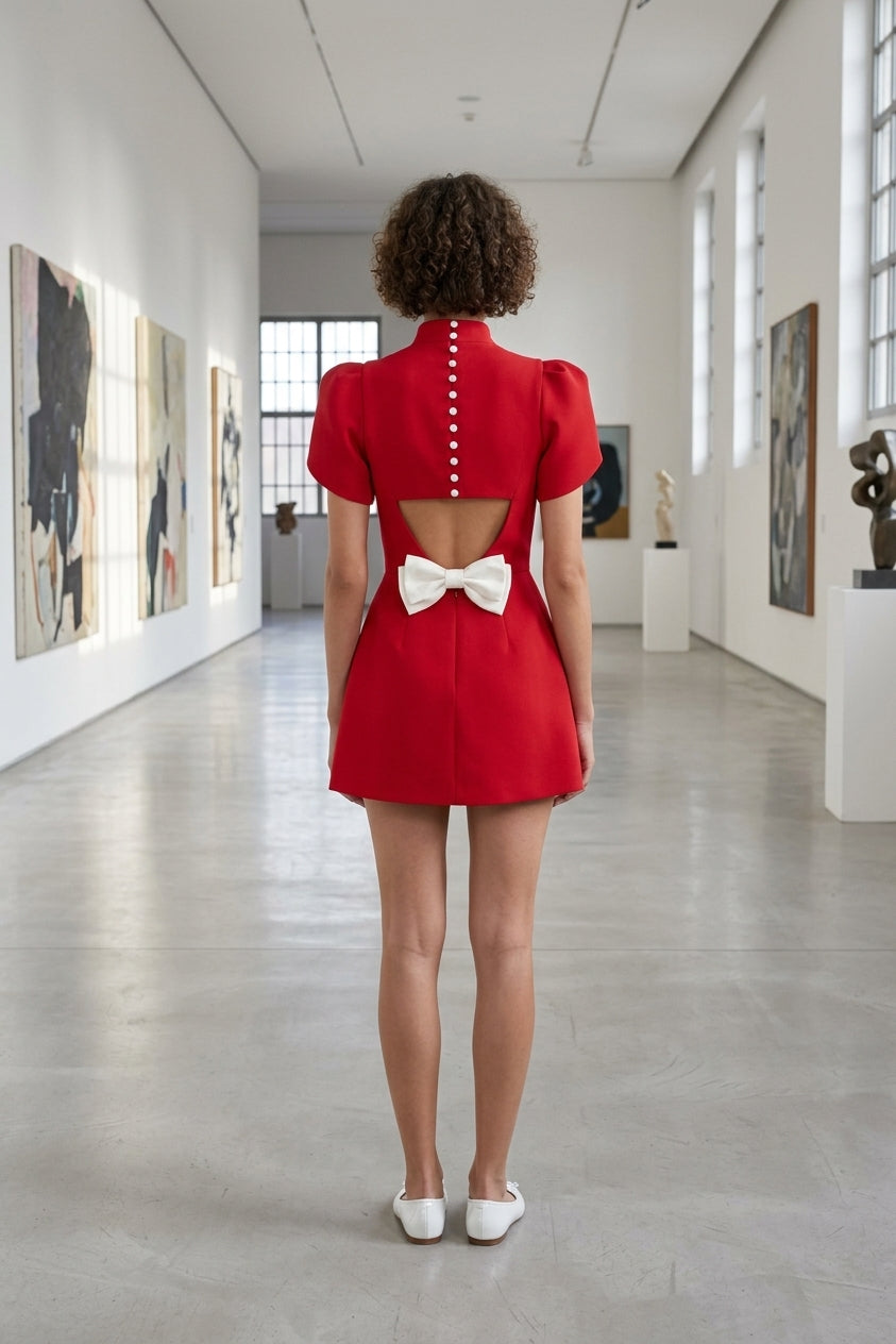 A woman viewed from behind wearing a bright red mini dress with puff sleeves, a high neckline, and a large white bow at the back of the open back. She is standing in a bright, modern art gallery with white walls, framed paintings, and a polished concrete floor.