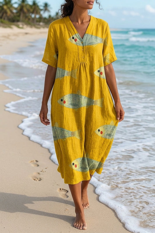 A woman wearing a yellow linen-like kaftan dress with a fish print walks along the shoreline of a sandy beach with turquoise water.