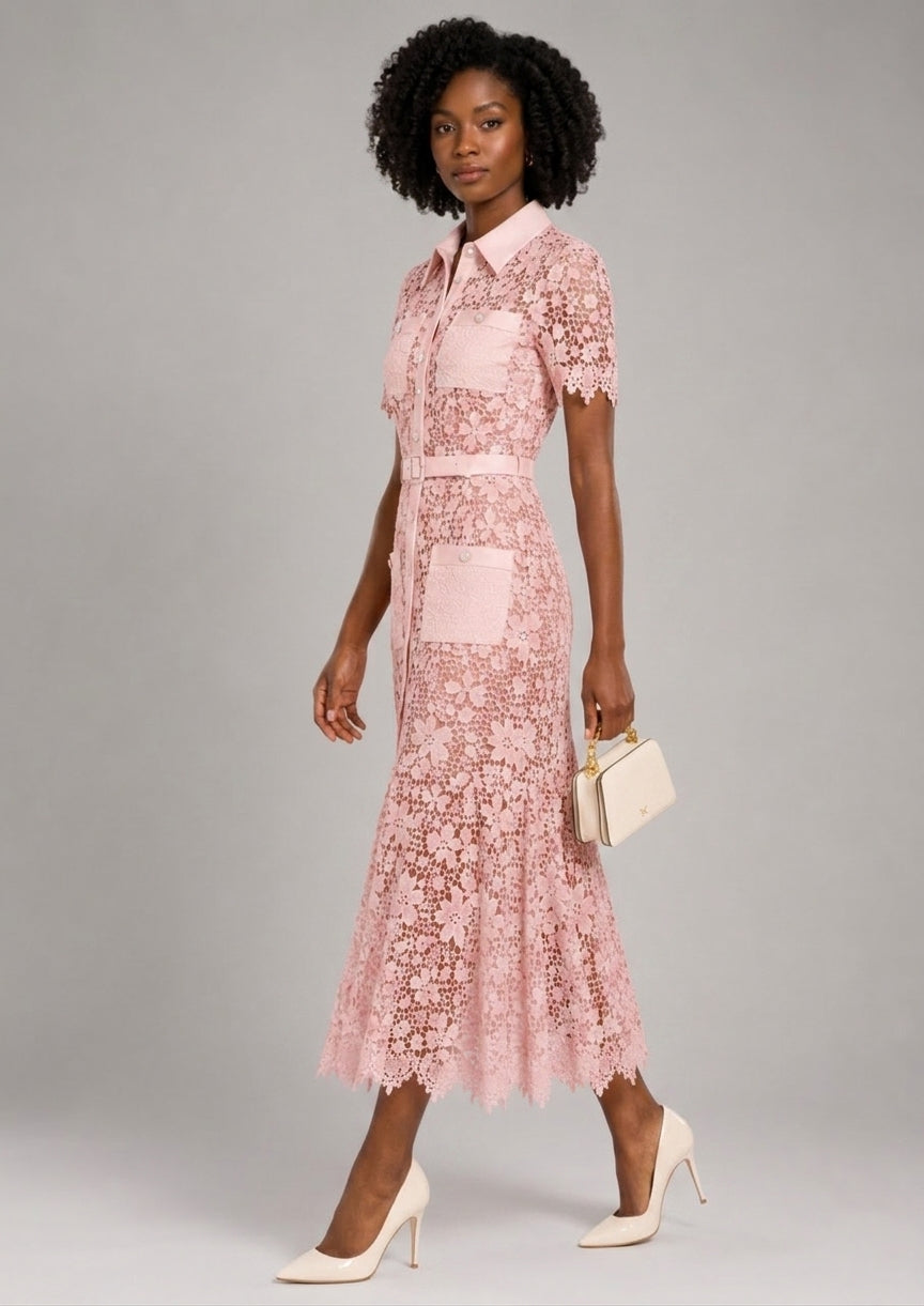 A woman models a light pink floral lace midi dress with short sleeves, a pointed collar, and a matching belt. She is holding a small cream clutch and wearing cream high heels, set against a solid gray studio background.