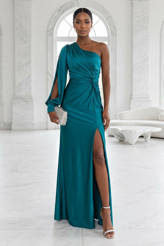 A woman models a floor-length teal green one-shoulder evening gown with a high thigh slit, draped waist detail, and a single sleeve with a crystal-embellished cuff, standing in a bright white marble hall.