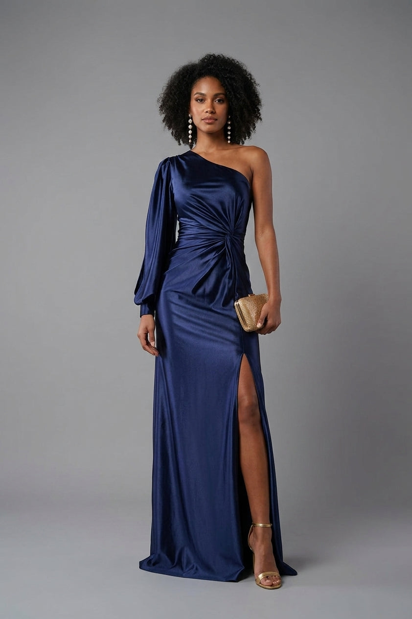 A woman modeling a floor-length navy blue satin gown with a one-shoulder design, long draped sleeve, gathered waist, and high front slit. She is standing against a plain gray studio background and holding a small metallic clutch.