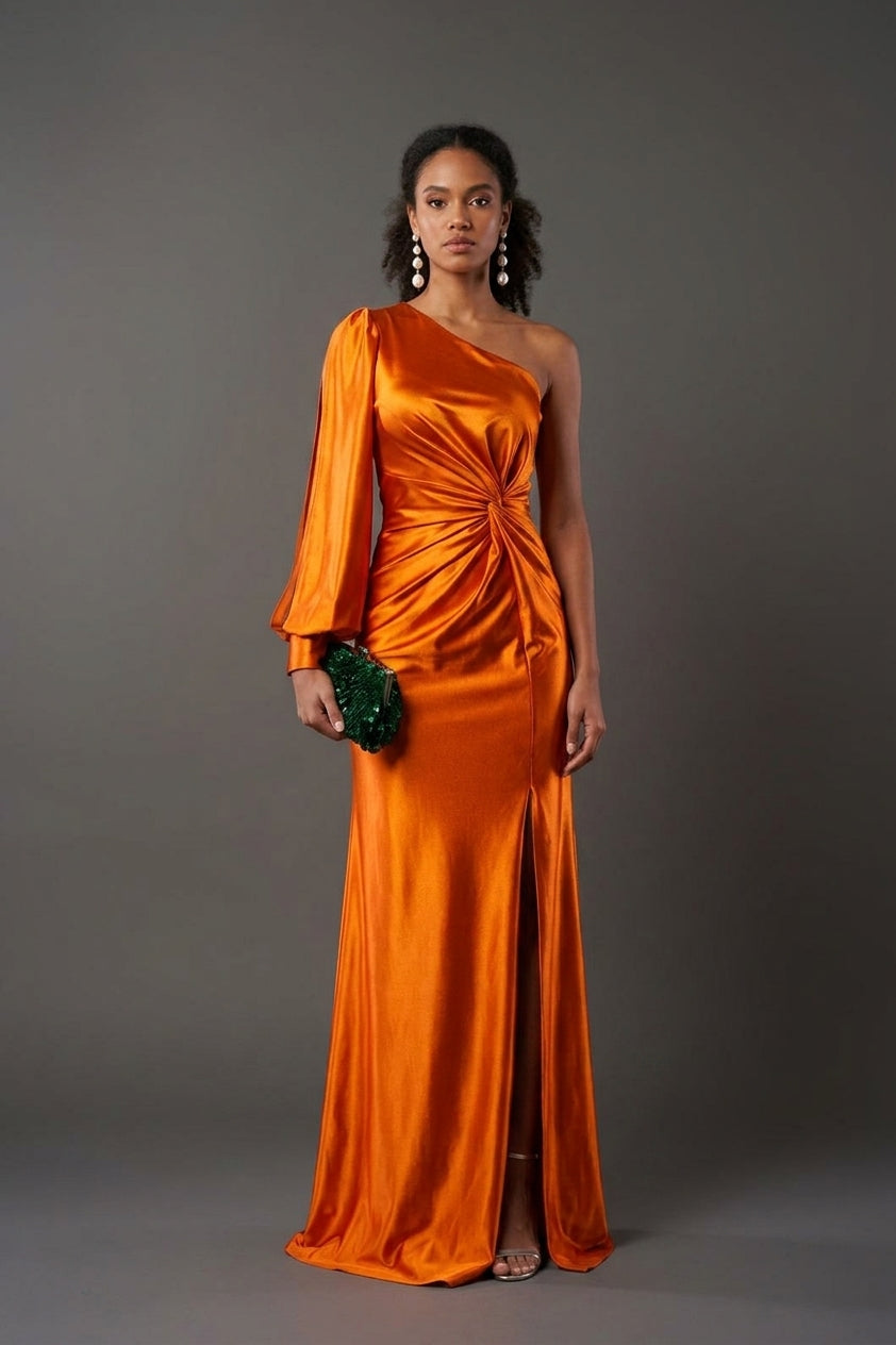 A woman modeling a floor-length, one-shoulder orange satin gown with a gathered waist and front slit, standing against a dark gray studio background and holding a small green clutch.