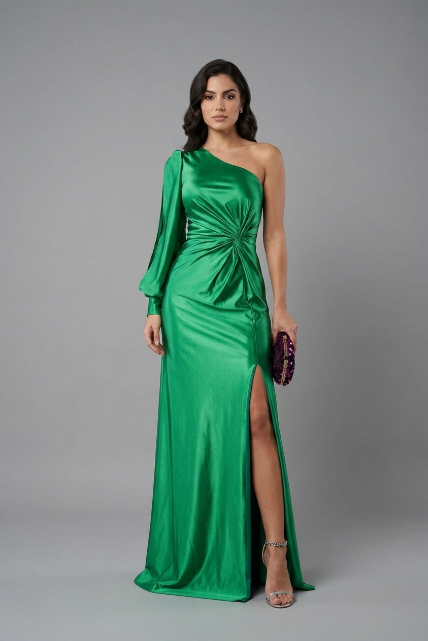A woman modeling a floor-length emerald green one-shoulder satin gown with a gathered waist, long sleeves, and a high front slit, standing against a plain gray studio background.