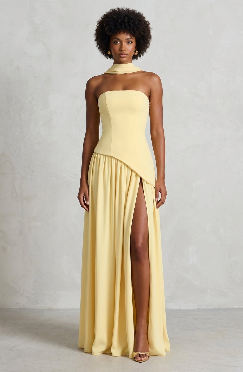 A woman modeling a pale yellow strapless maxi dress with a high neck, structured bodice, and a high thigh slit, set against a textured light gray studio background.