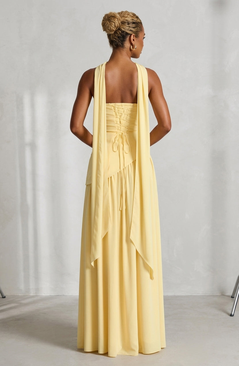 A woman shown from the back wearing a pale yellow sleeveless maxi dress with a draped shawl-style back overlay, photographed in a bright studio setting.