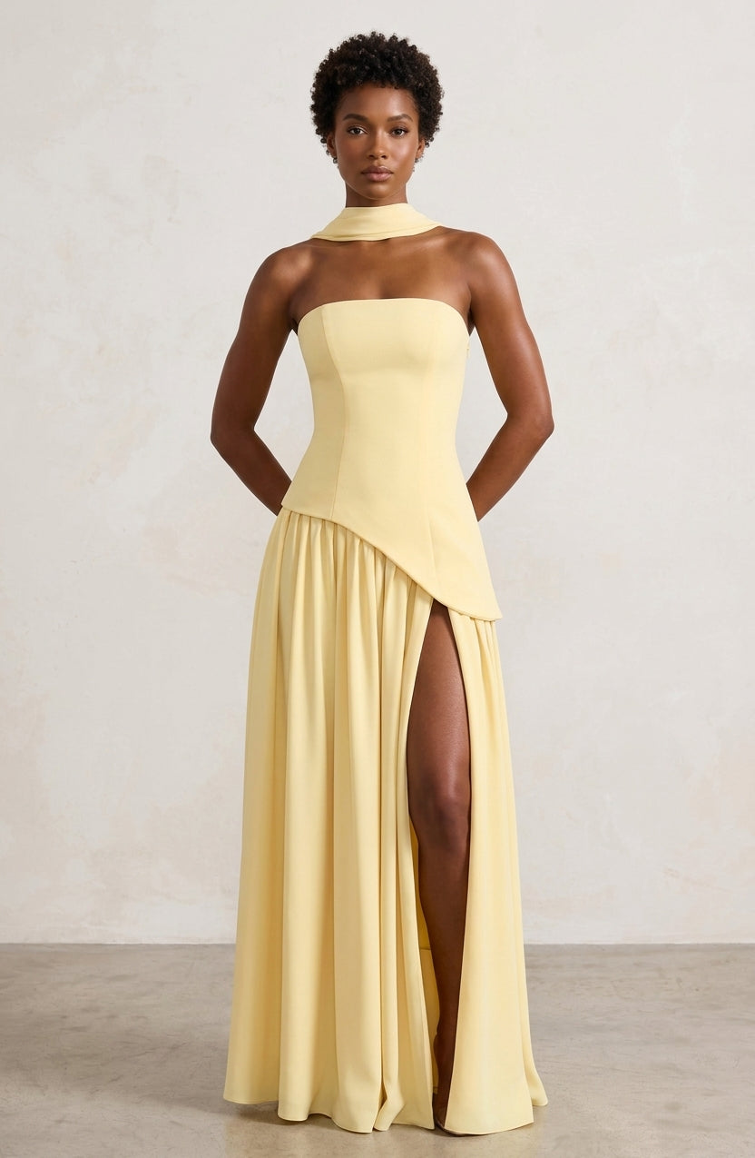 A model wearing a pale yellow strapless gown with a high neck, structured bodice, and a high thigh slit, standing against a neutral textured studio wall.