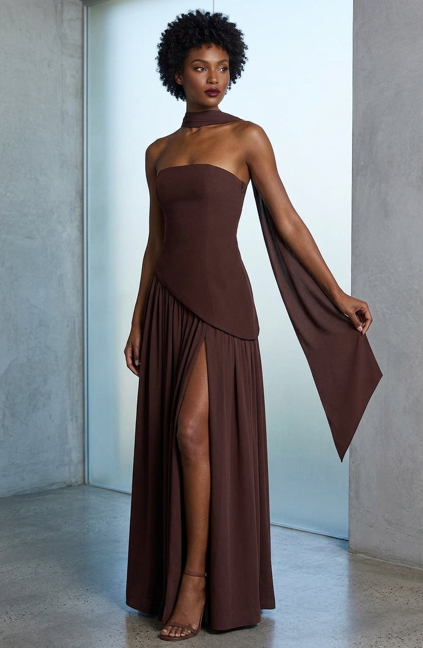 A woman models a floor-length brown strapless gown with a high slit, draped cape-like sleeve, and choker neckline in a minimalist studio setting.