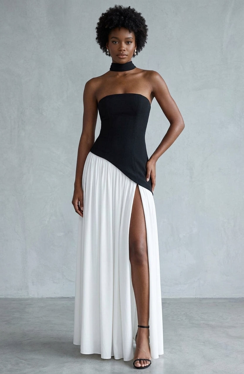 A model wearing a strapless black and white maxi dress with a high thigh slit, standing against a textured gray studio backdrop.