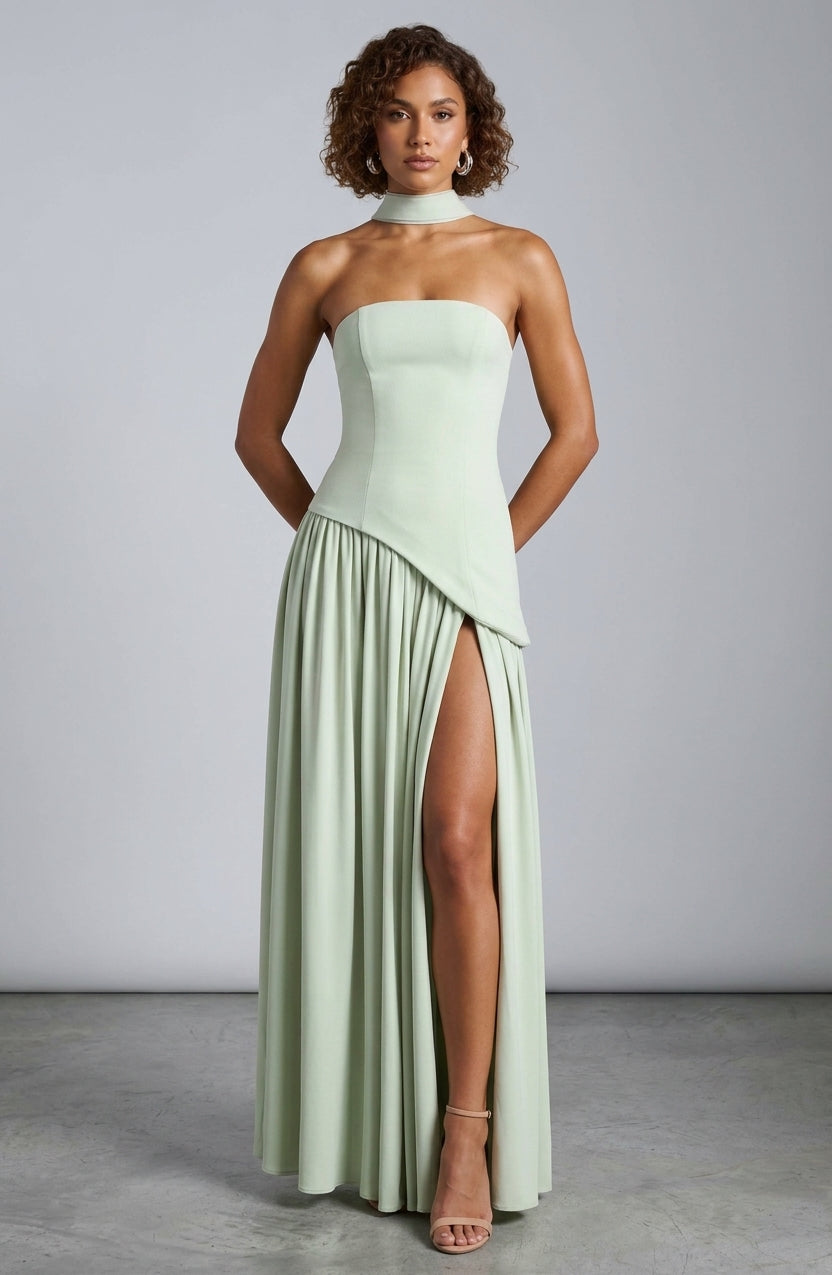 A woman models a strapless mint green maxi dress with a high neck, structured bodice, draped asymmetrical overlay, and a high thigh slit, set against a neutral gray studio background.