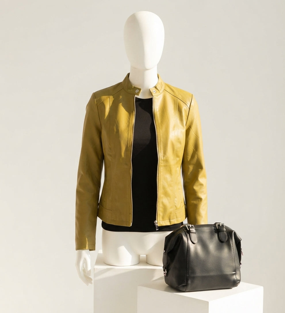 A mustard yellow leather jacket displayed on a white mannequin torso, styled over a black top, with a black leather handbag resting on a white pedestal against a light gray studio background.