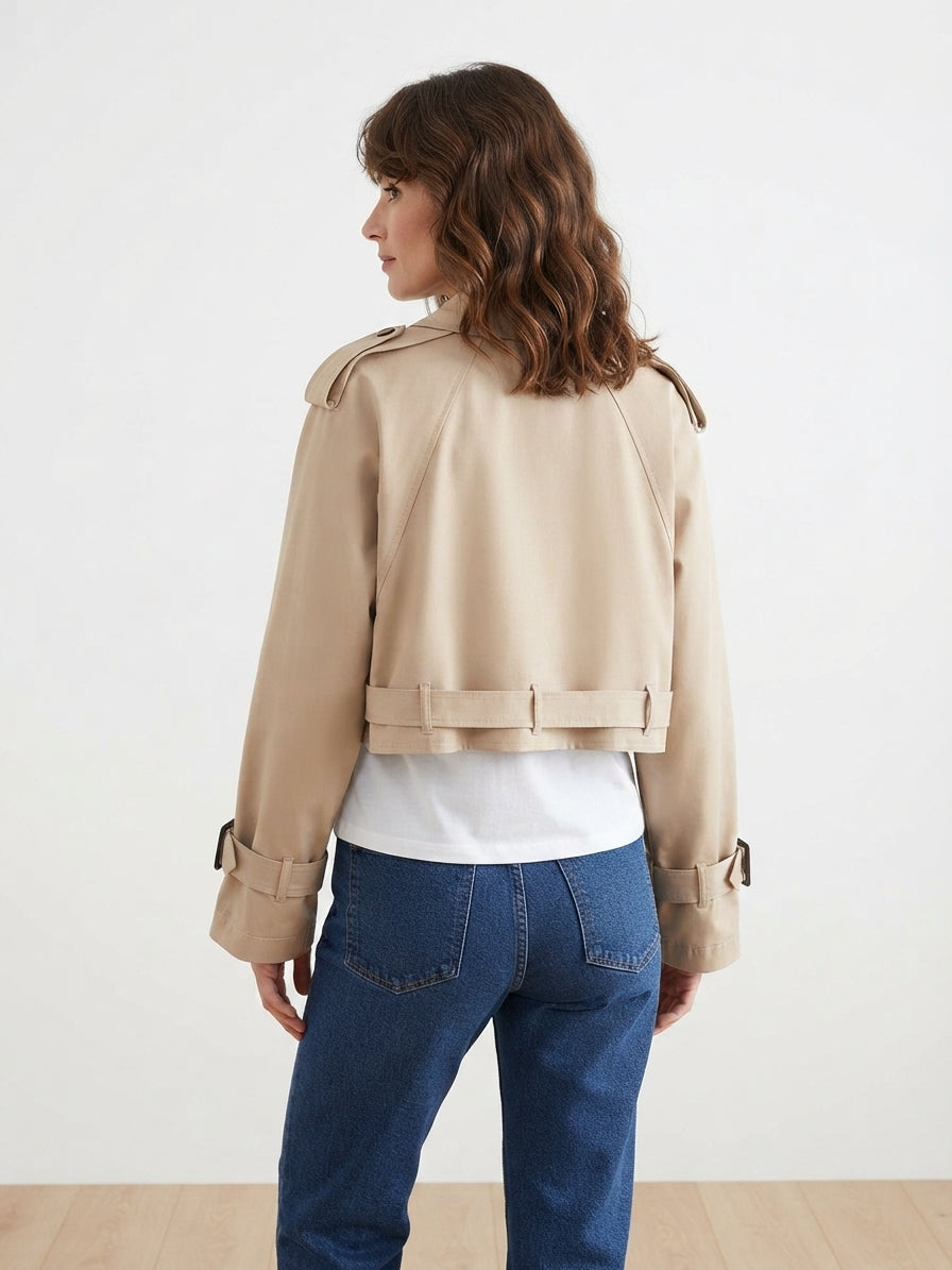 A woman shown from the back wearing a cropped beige trench-style jacket over a white top and blue jeans, photographed in a clean studio setting.