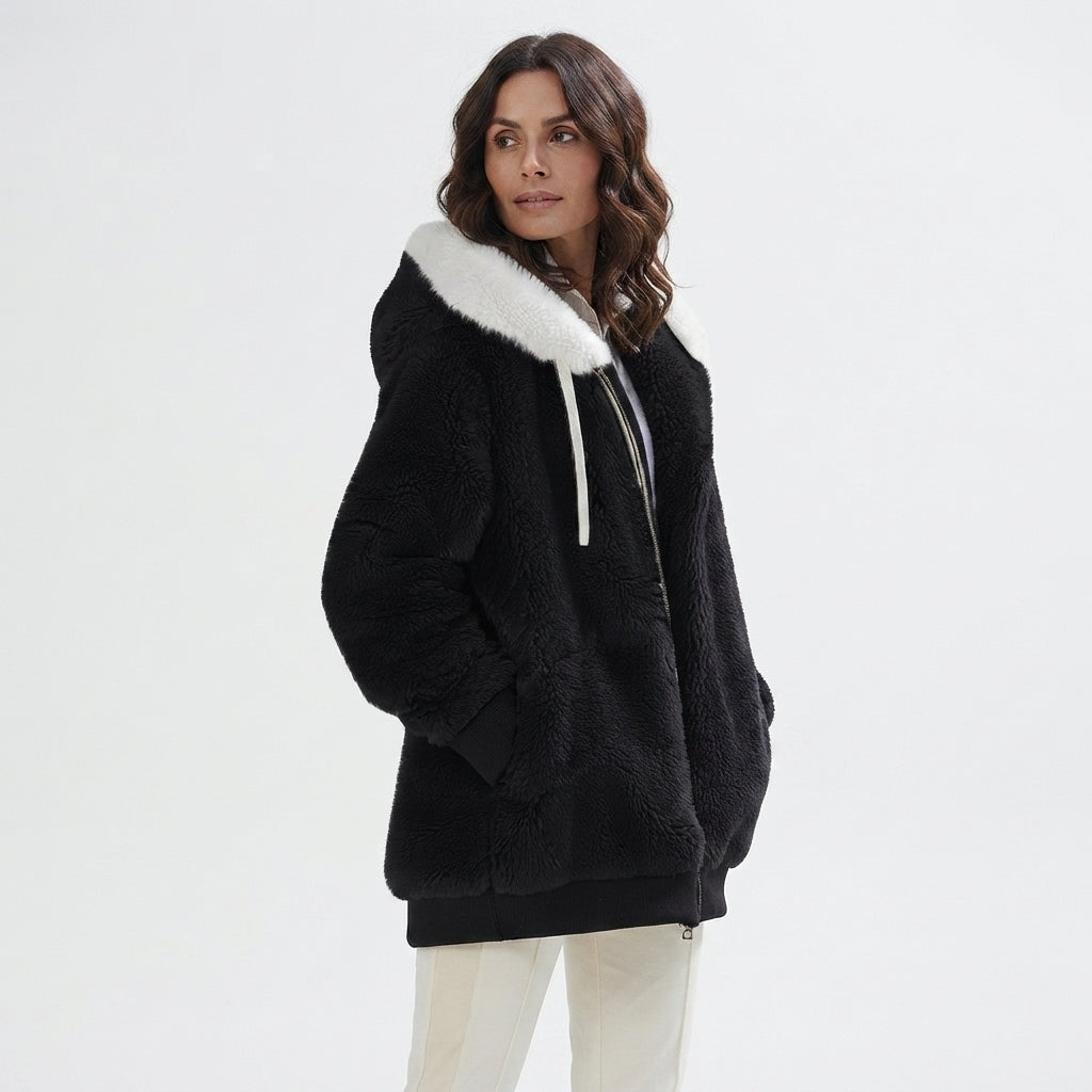 A woman wearing a black sherpa fleece hooded jacket with a contrasting white faux-fur collar and white drawstrings, paired with white pants, against a plain white studio background.