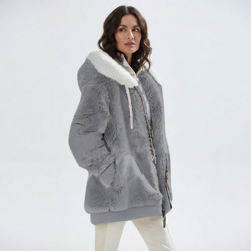 A woman wearing a gray plush faux-fur hooded jacket with a white faux-fur collar and white drawstrings, paired with off-white pants, against a plain light gray studio background.