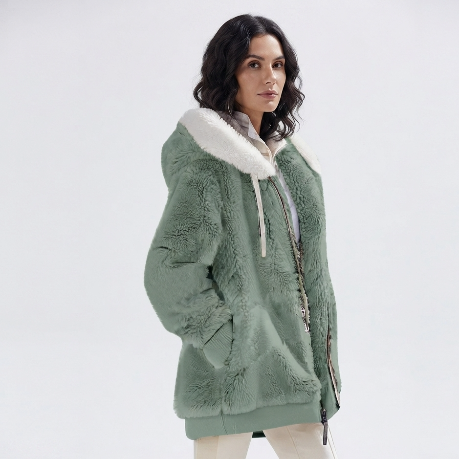 A woman wearing a sage green plush fleece hooded jacket with a white faux-fur collar, standing against a plain light gray studio background.