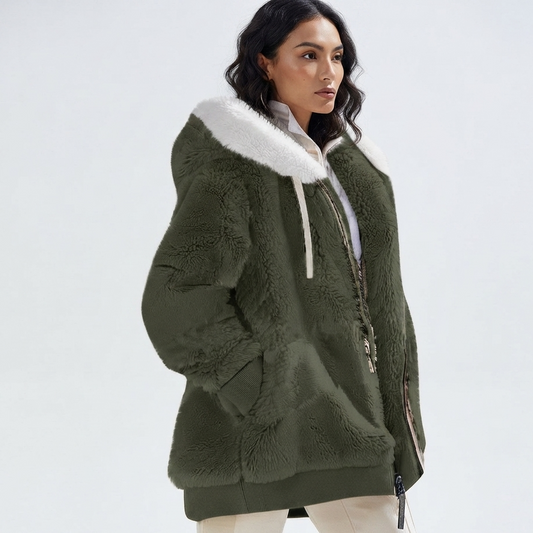 A woman wearing an olive green sherpa fleece jacket with a white faux-fur trimmed hood and white drawstrings, paired with white pants, against a plain light gray studio background.